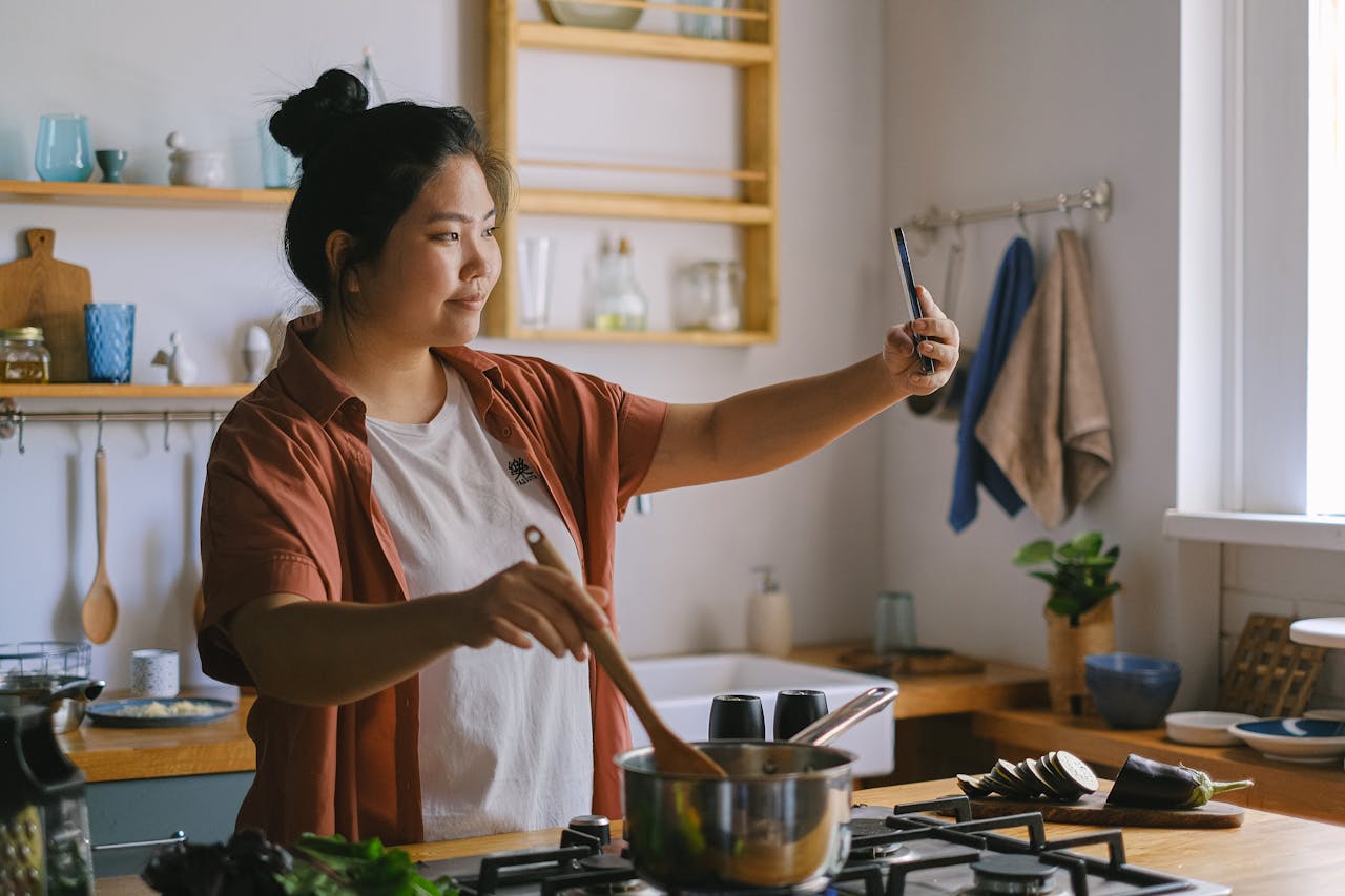 gallery-02 Woman cooking in kitchen and taking selfie, capturing the moment.