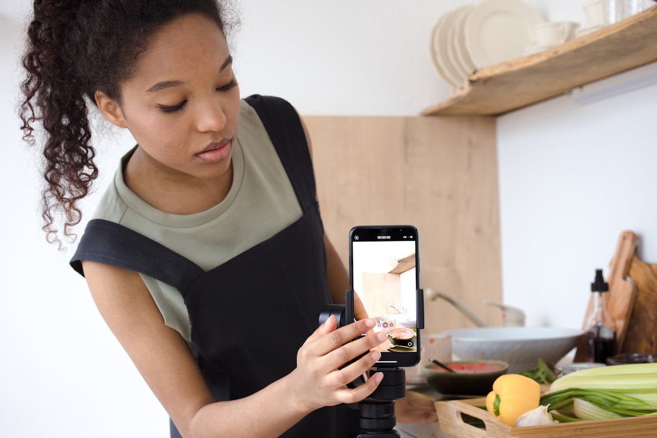 gallery-04 A young woman filming a cooking tutorial in her kitchen using a smartphone.